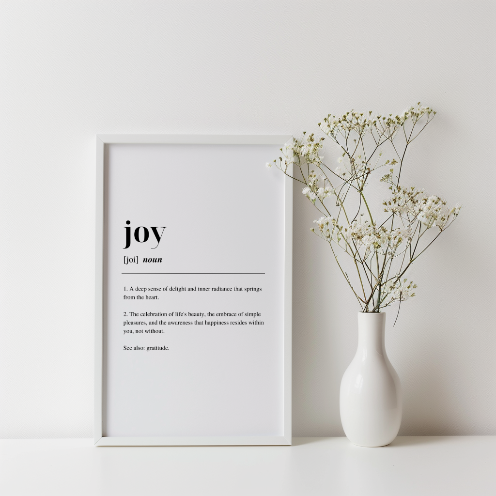 White framed poster with inspirational definition art text for the word and meaning of Joy. there is a plant in a vase to the right of the frame. the background wall is white. 