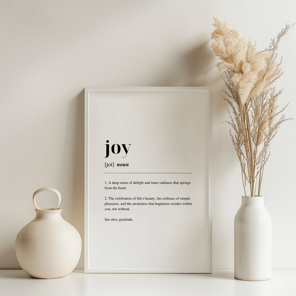 Dark framed poster with inspirational definition art text for the word and meaning of Joy. the frame is surrounded by plants against a white wall with a dried plant  in a vase to the right.