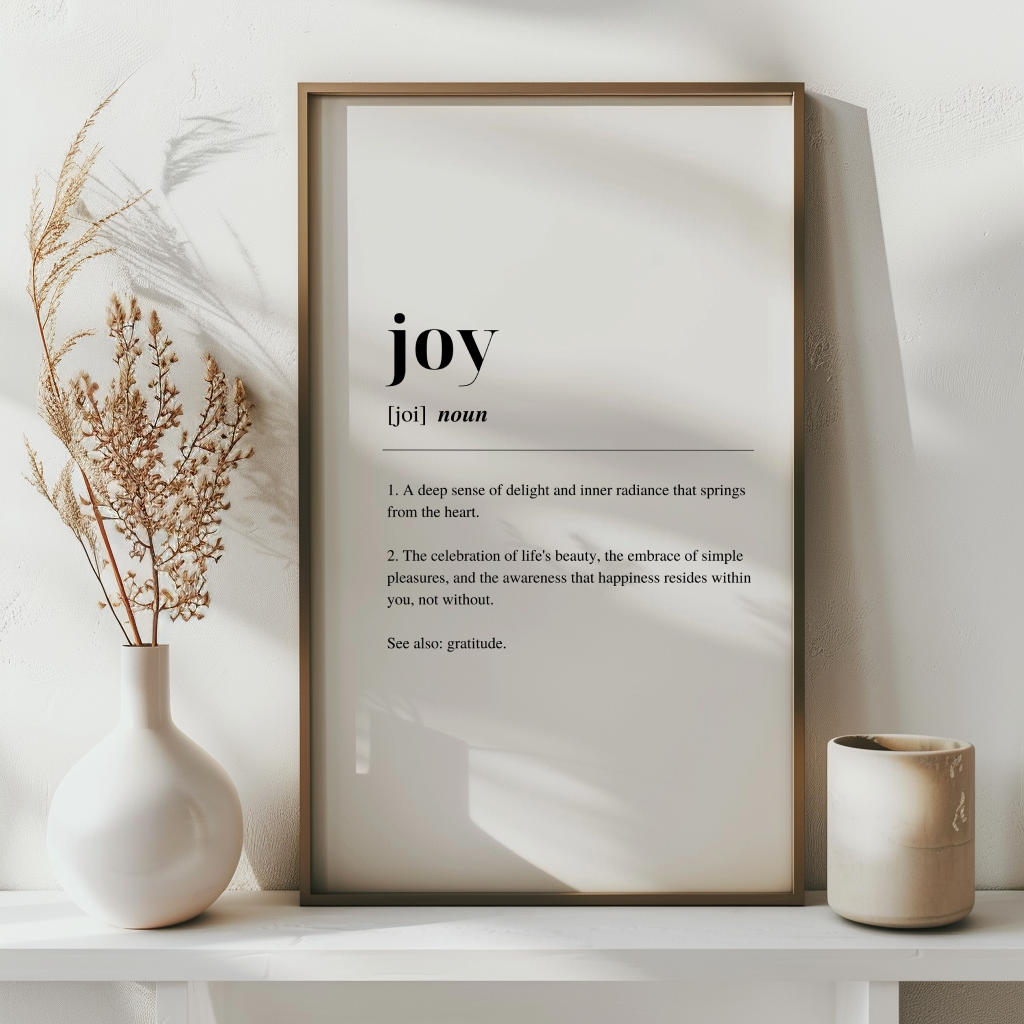 Dark framed poster with inspirational definition art text for the word and meaning of Joy. the frame is surrounded by plants against a white wall with a dried plant  in a vase to the left.