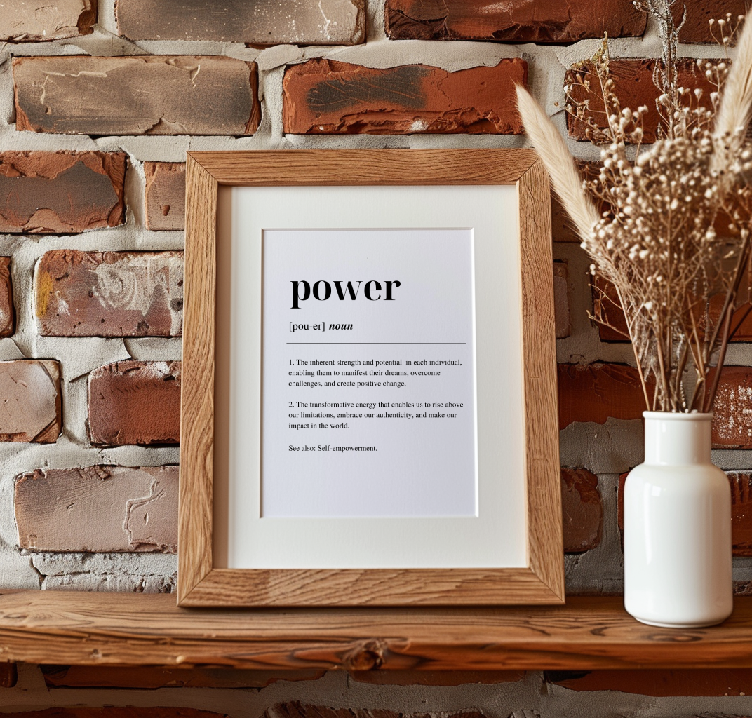 A digital art print with the word 'POWER' written in black capital letters, defined as 'the ability to transform weakness into strength and to face challenges with determination'. The print is framed in a wooden frame, displayed a brick wall, on a mantlpiece with a vase with a dried plant  to the right.